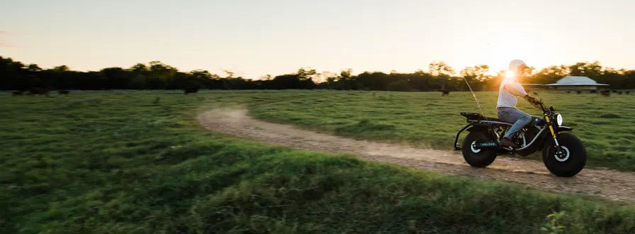 Person riding a motorcycle on a dirt path in a grassy field at sunset.
